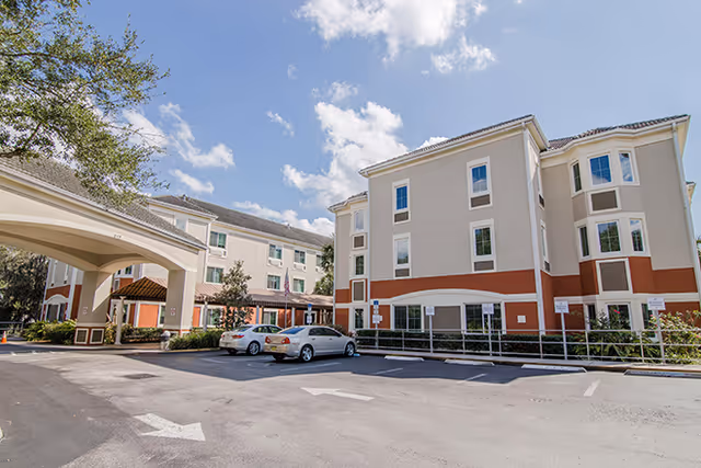 Exterior view of a multi-story senior living facility building with beige and brown walls, several windows, and a covered entrance. There are a few parked cars in front of the building under a bright blue sky with some clouds.
