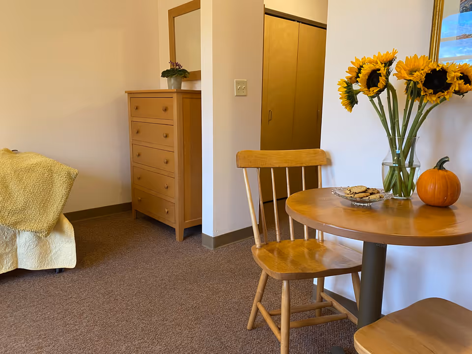 A cozy bedroom corner with a bed, wooden dresser, and a small round table topped with sunflowers and a pumpkin.