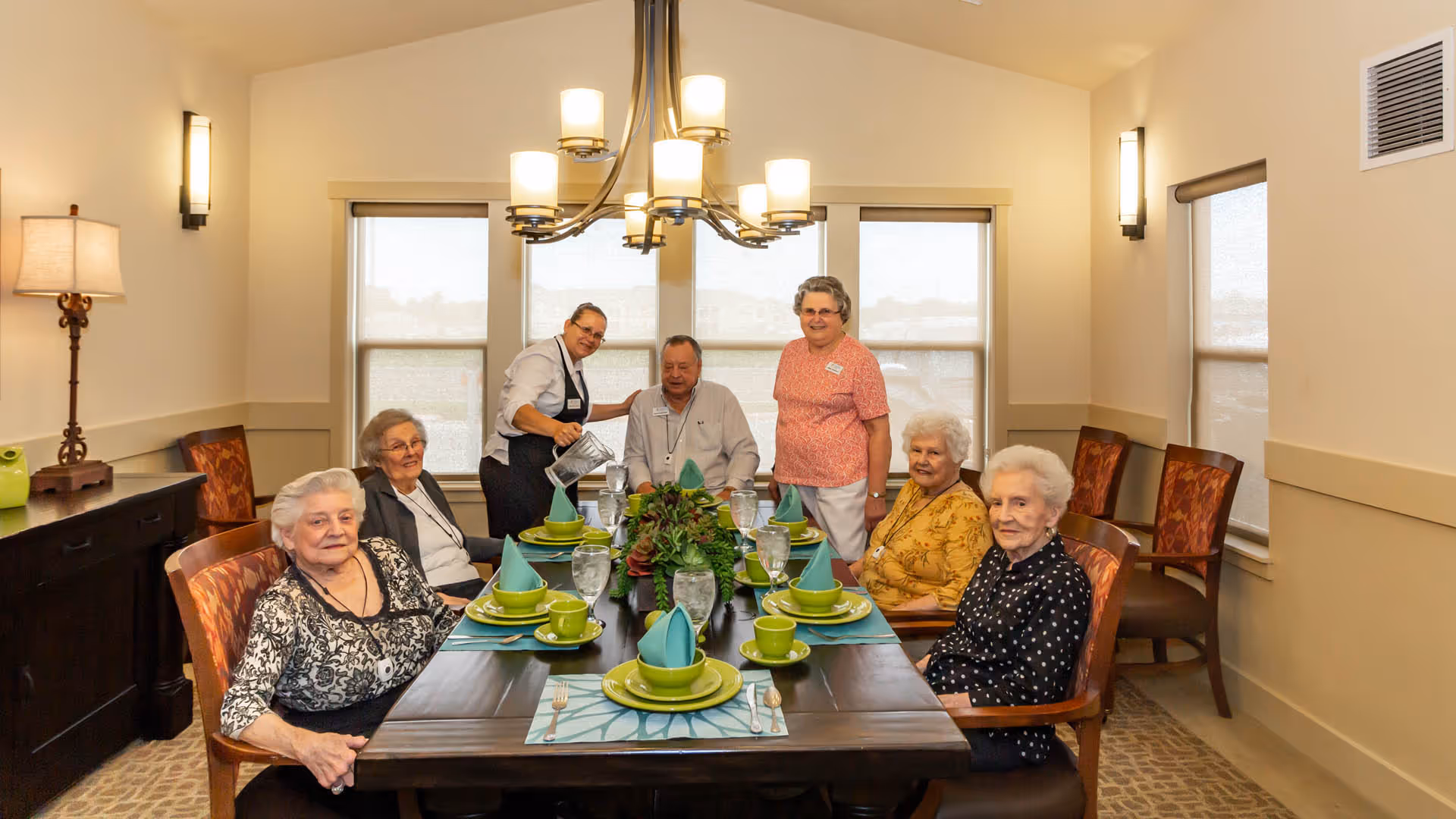 A group of elderly people seated around a dining table set with green plates, cups, and blue napkins in a well-lit dining room. A staff member is pouring water into a glass while another staff member stands nearby smiling. The room has large windows with shades, a chandelier, and a sideboard with a lamp.