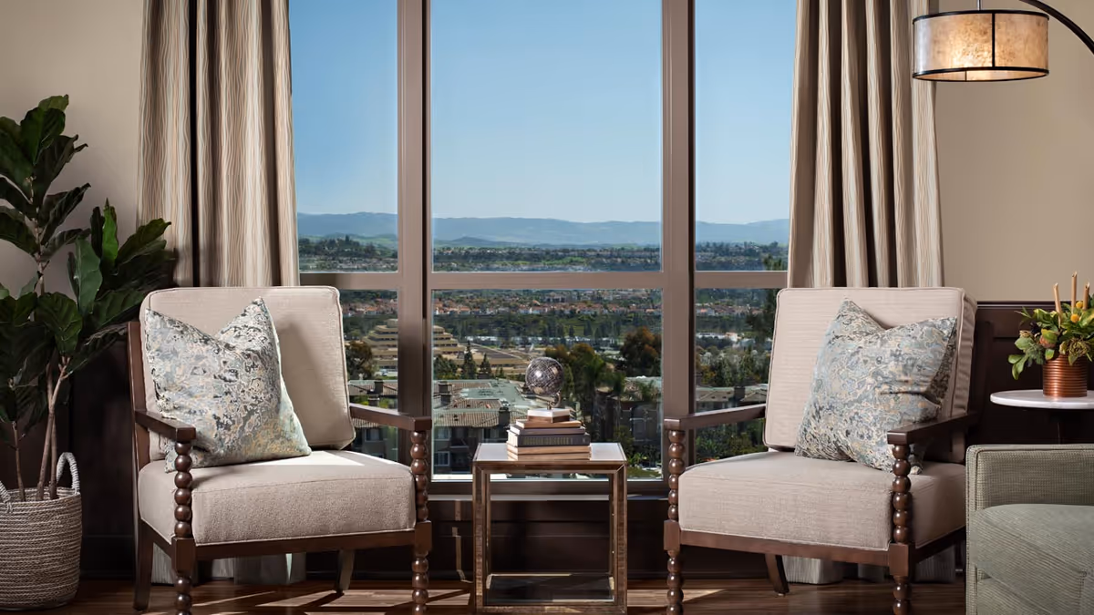 Two beige armchairs with patterned cushions are positioned on either side of a small glass-top table holding a stack of books and a decorative globe. Behind the chairs is a large window offering a clear view of a cityscape with buildings, trees, and distant hills under a blue sky. A tall green plant in a woven basket is on the left, and a round side table with a floral arrangement and a hanging lamp are on the right.