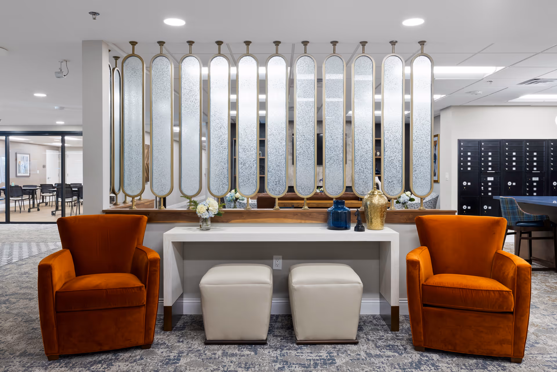 A modern senior living facility common area with two orange armchairs on either side of a white console table. The table has decorative vases and flowers on top, with two white ottomans underneath. Behind the table is a decorative partition with vertical frosted glass panels. In the background, there are black mailboxes and a seating area with chairs and tables.