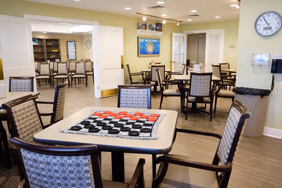 A well-lit common area in a senior living facility with multiple tables and chairs. One table in the foreground has a checkers game set up with red and black pieces. The room has light green walls, a clock on the wall, and a trash can near the corner. In the background, there are more chairs and tables, some with decorative items and artwork on the walls.