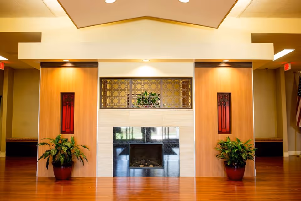 A modern indoor fireplace set in a beige and black tiled wall with decorative metal grille above it. The fireplace is flanked by two wooden panels each with a vertical red stained glass window. Two potted green plants are placed on the floor in front of the wooden panels. The floor is wooden and the ceiling has recessed lighting.