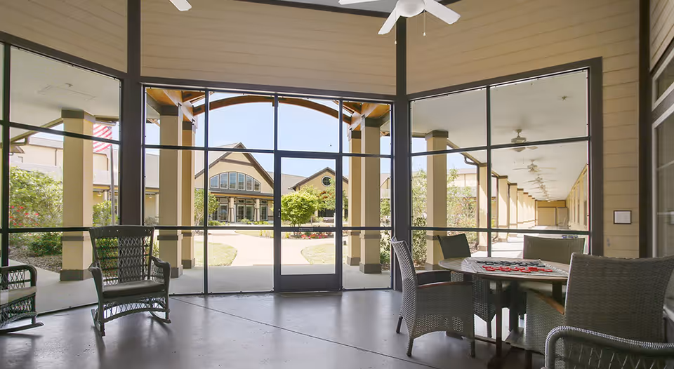 A covered outdoor seating area with wicker chairs and a round table with a checkerboard game on it. The area is enclosed with large glass windows and doors, showing a view of a walkway, garden, and a building with a peaked roof in the background.