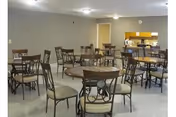 Communal dining room with several round tables and padded chairs and a serving counter in the background.