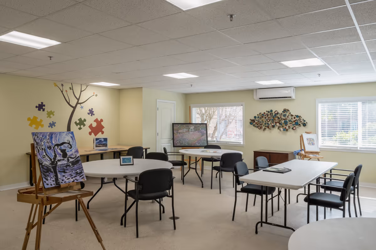 A well-lit activity room with several tables and chairs arranged for group activities. The room features artwork on easels, colorful puzzle piece wall decals, a decorative wall sculpture, and large windows with blinds letting in natural light. The walls are painted a soft yellow, and the ceiling has recessed lighting panels.