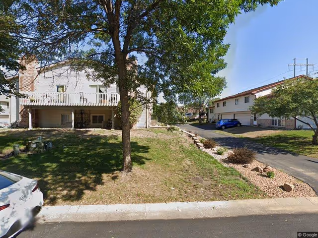 Exterior view of a residential area with two multi-unit buildings, a tree in the foreground, a driveway with parked cars, and a grassy lawn with some landscaping under a clear blue sky.