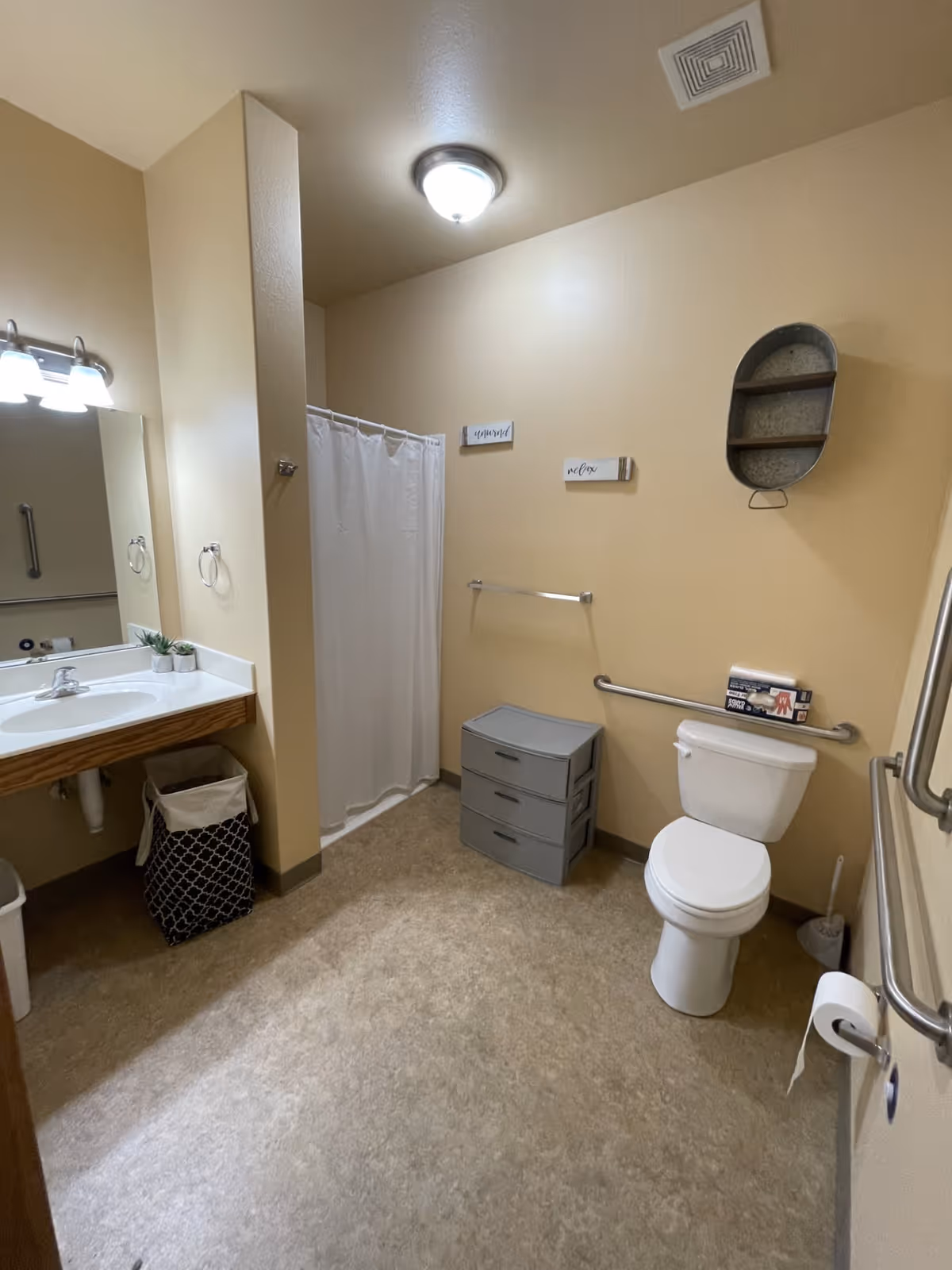 A clean bathroom with beige walls and flooring, featuring a white toilet with a grab bar, a gray three-drawer storage unit, a shower with a white curtain, a sink with a mirror and light fixture above it, a laundry basket, and small decorative signs on the wall.