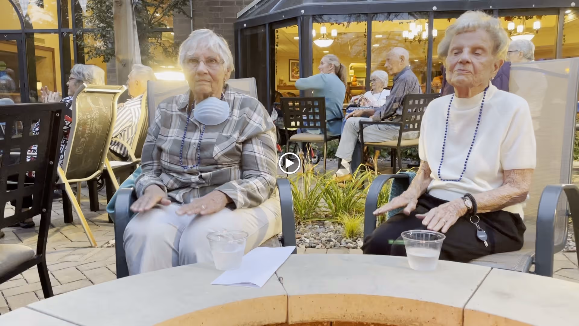Two elderly women sitting outdoors on patio chairs around a circular stone table with plastic cups of water. Behind them, other seniors are seated and conversing near a building with large windows and warm indoor lighting.