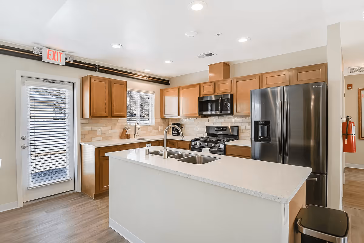 Modern kitchen with wooden cabinets, a large island with a double sink, stainless steel refrigerator, stove, and microwave. There is a door with blinds and a window above the sink allowing natural light into the space.