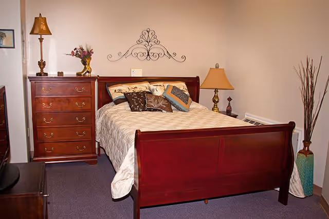 A bedroom with a wooden bed frame featuring a beige quilt and multiple decorative pillows. To the left of the bed is a wooden dresser with a lamp and a vase of flowers on top. On the right side of the bed is a small table with a lamp and decorative items. A tall vase with long sticks is placed in the corner of the room. The walls are light-colored and there is a decorative metal wall hanging above the bed.