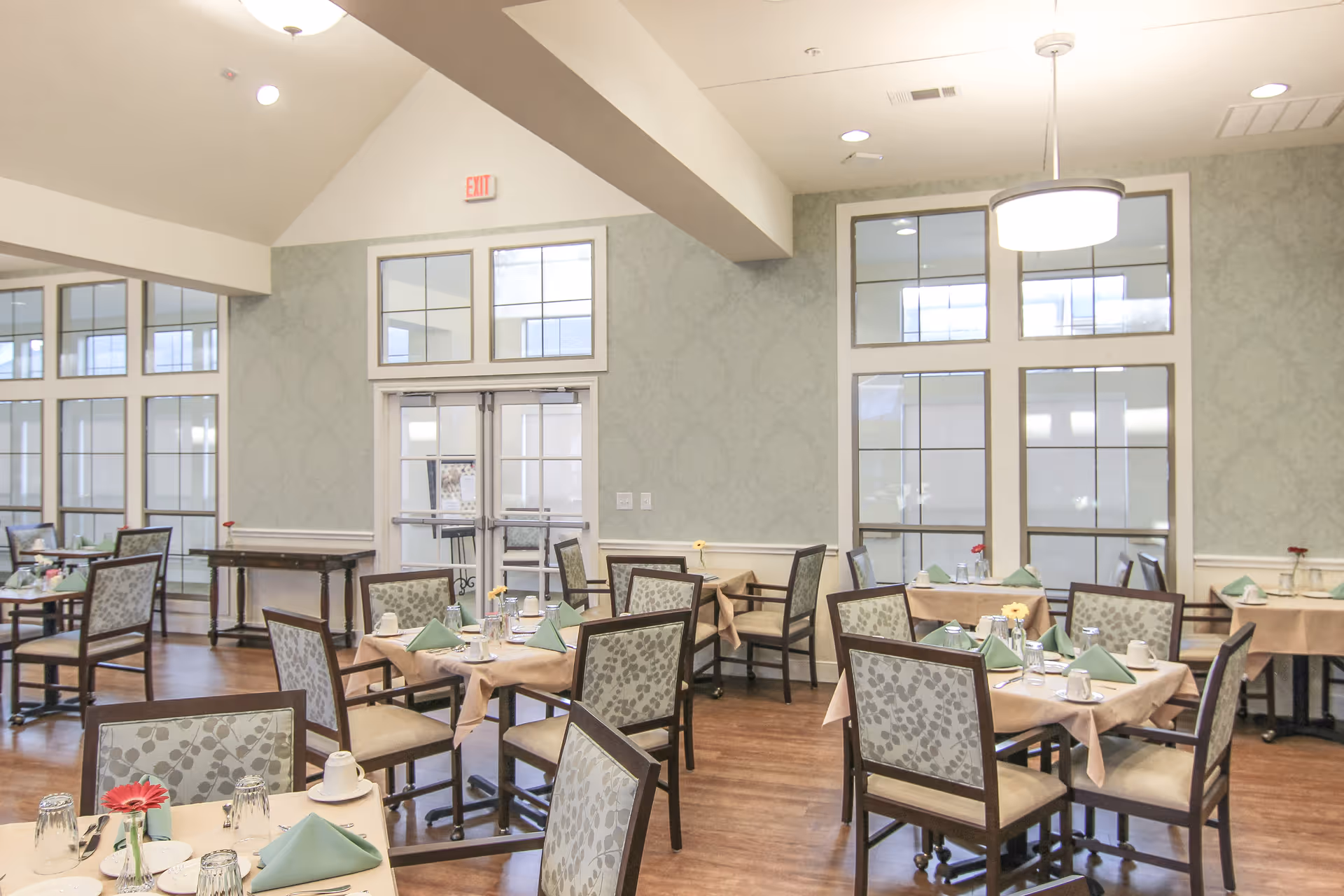 A bright dining room with several tables covered with beige tablecloths, each set with green folded napkins, cups, glasses, and silverware. The room has large windows with white frames, light green patterned wallpaper, wooden flooring, and a modern ceiling light fixture.