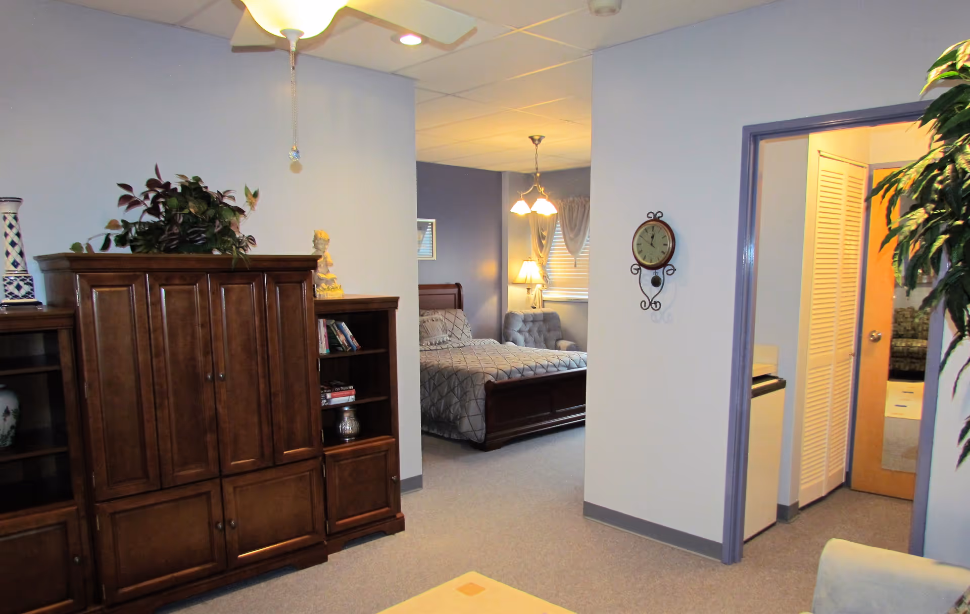 Interior view of a senior living facility room at St. Dominic Village showing a wooden entertainment cabinet with decorative items on top, a bedroom with a bed and a chair visible through an open doorway, a wall clock, and part of a living area with a plant and a ceiling fan.
