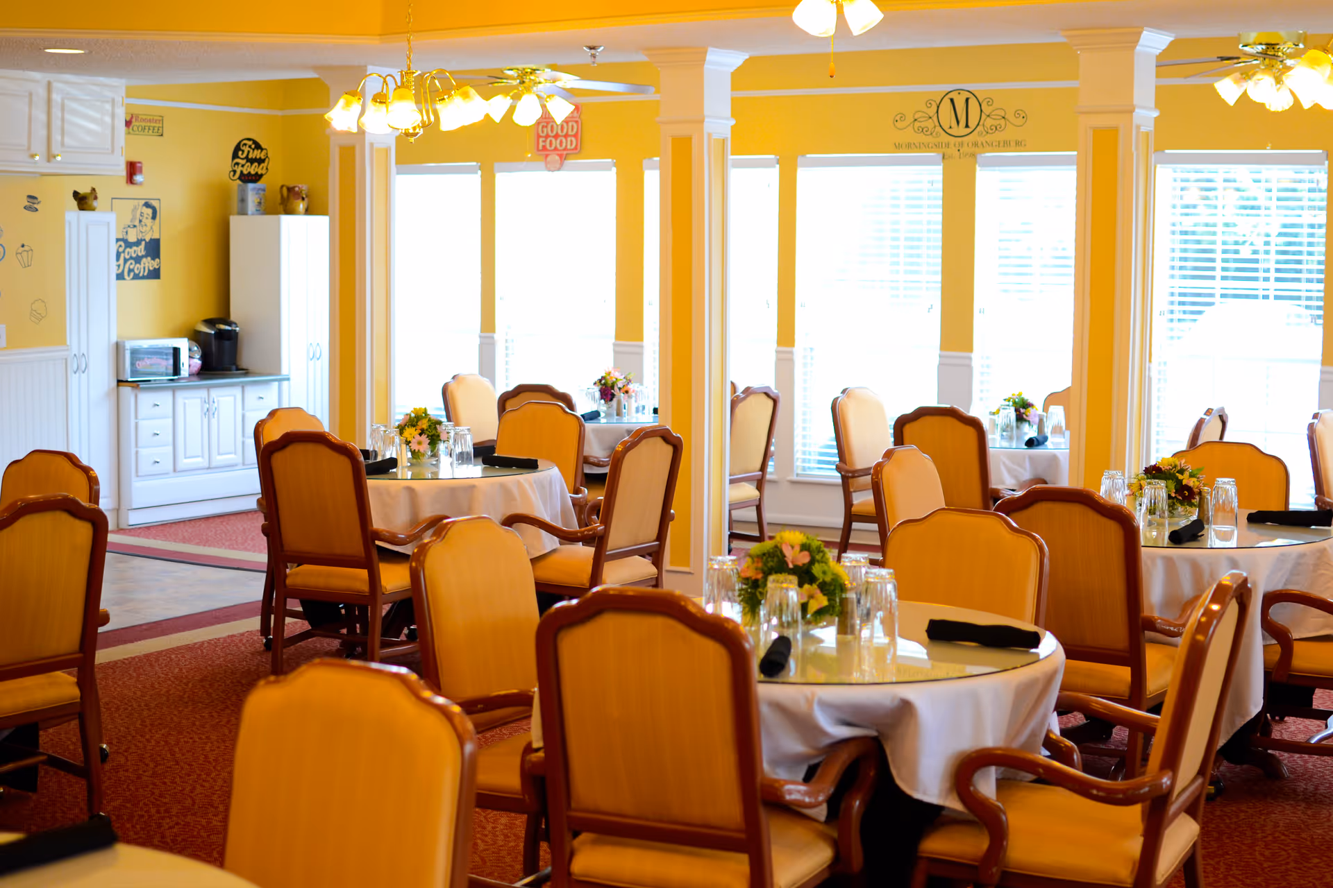 A bright dining room with yellow walls and multiple round tables covered with white tablecloths. Each table is set with glasses, black napkins, and small floral centerpieces. The room has wooden chairs with cushioned seats and backs. Large windows with white blinds allow natural light to fill the space. Ceiling fans with lights hang from the ceiling, and there is a small kitchenette area with a microwave and coffee maker in the background.