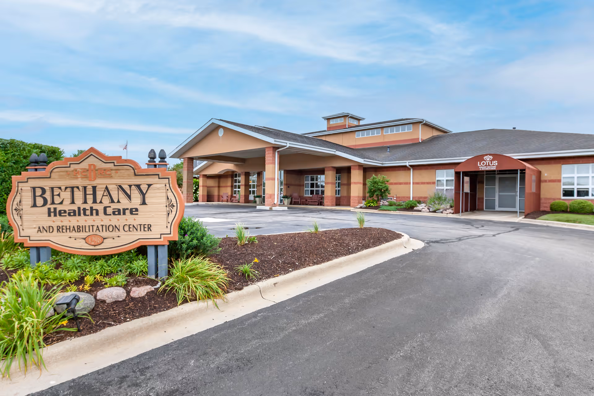 Exterior view of Bethany Health Care and Rehabilitation Center building with a large wooden sign in the foreground displaying the facility name. The building has a covered entrance with columns and a red awning labeled 'Lotus'. The surrounding area includes landscaped greenery and a paved driveway.