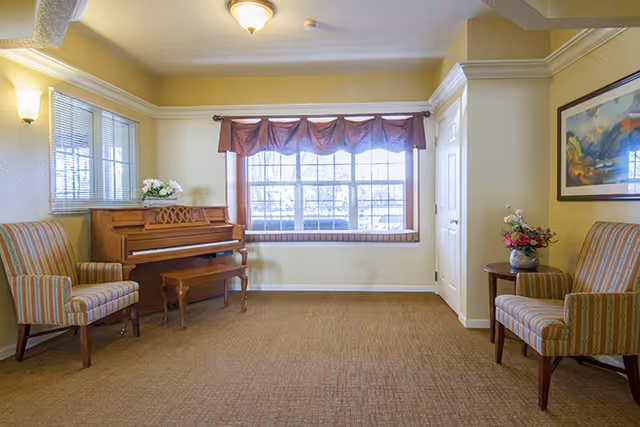 A cozy room with beige walls and carpet, featuring a wooden piano with a matching bench on the left side, two striped armchairs on either side of the room, a small round table with a flower vase, a large window with a brown valance, and a framed landscape painting on the right wall.