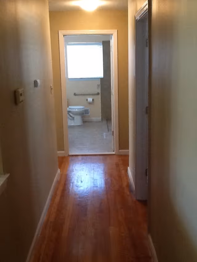 View down a narrow hallway with wooden flooring leading to a bathroom. The bathroom has a toilet, a grab bar, a toilet paper holder, and a window with blinds. The walls in the hallway are painted beige.