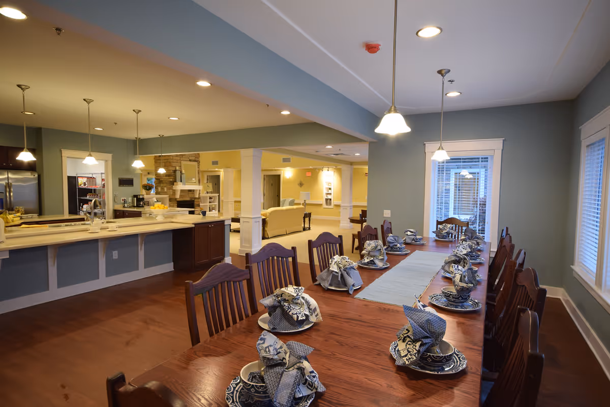 A spacious dining area in a senior living facility featuring a long wooden table set with blue and white patterned dishes and folded napkins. The room has wooden chairs, pendant lights hanging from the ceiling, and large windows with white blinds. In the background, there is an open kitchen area with a countertop, stainless steel refrigerator, and a cozy living space with beige sofas and a stone fireplace.