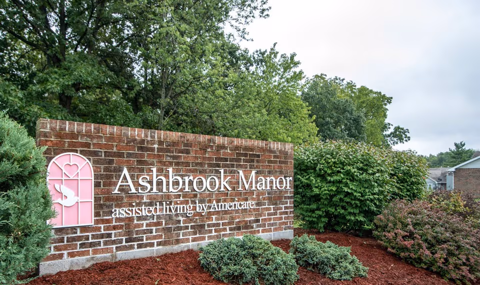 Brick entrance sign reading 'Ashbrook Manor assisted living by Americare' surrounded by landscaped bushes and trees.