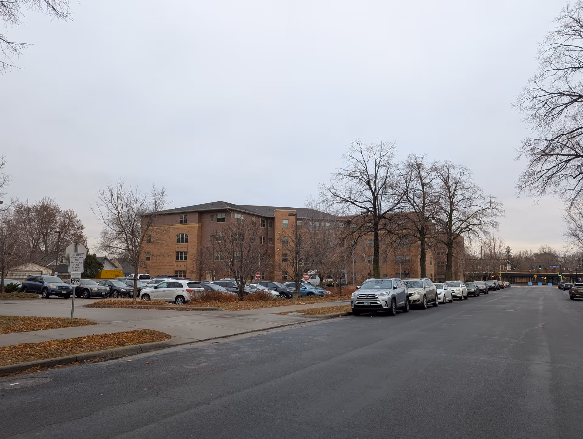 Exterior view of a multi-story brick building with a parking lot in front, several parked cars along the street, and leafless trees under an overcast sky.