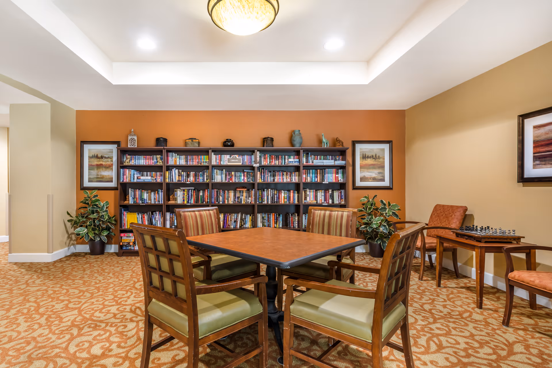 A cozy reading and game area in a senior living facility featuring a square wooden table surrounded by four cushioned chairs with striped upholstery. Behind the table is a large bookshelf filled with books and decorative items. The room has warm-toned walls with framed artwork, potted plants, and a small side table with a chessboard and two chairs. The ceiling has recessed lighting and a central light fixture.