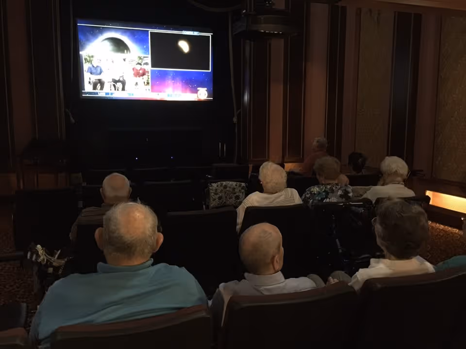 A group of elderly people seated in a dimly lit room watching a large screen television mounted on the wall. The room has dark wood paneling and patterned carpet, resembling a small theater or media room.