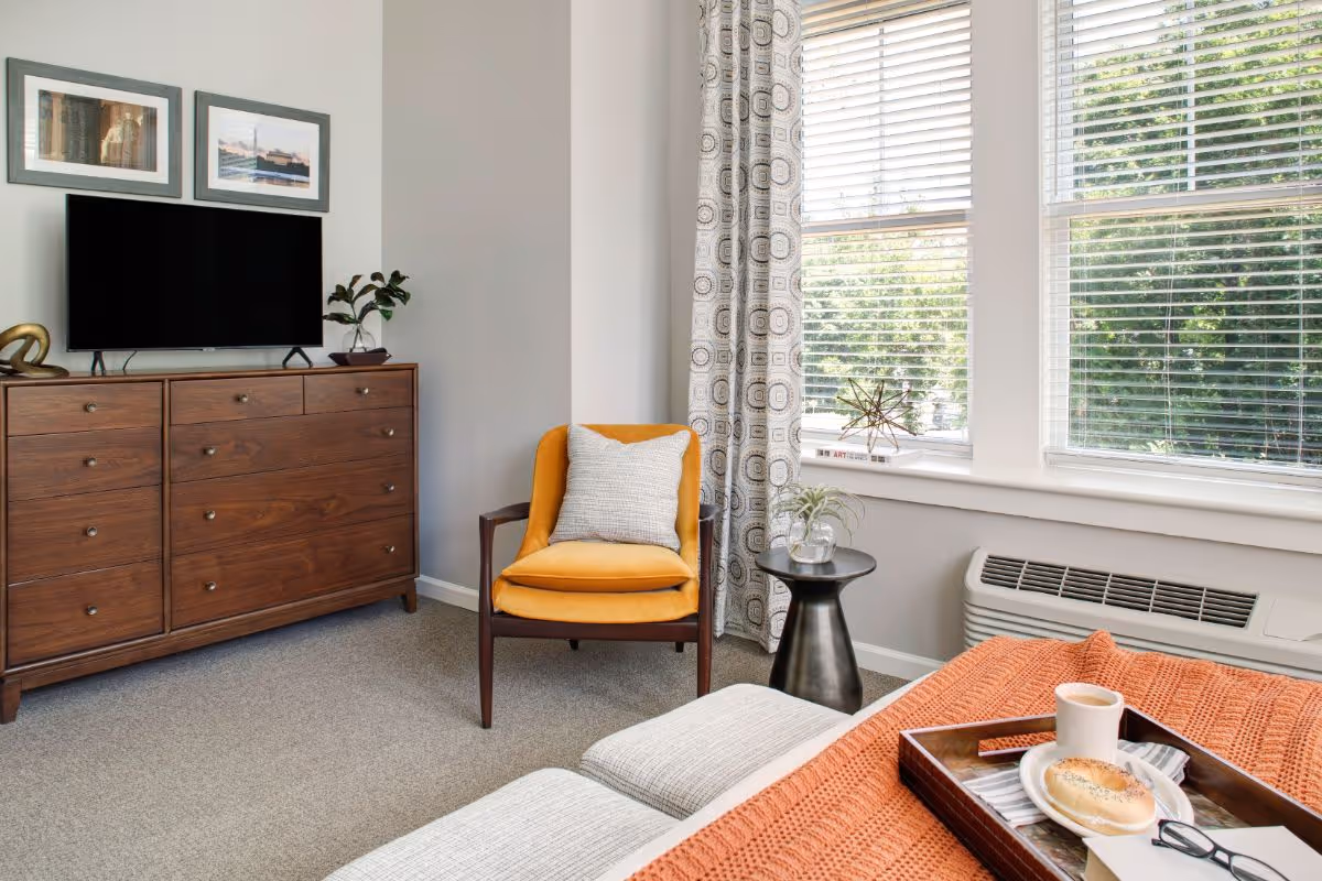 A cozy bedroom corner with a wooden dresser holding a flat-screen TV and decorative items, a mustard yellow armchair with a white pillow, a small black side table with a plant, and large windows with patterned curtains letting in natural light. In the foreground, part of a bed with an orange knitted blanket and a tray holding a bagel, coffee cup, and glasses is visible.
