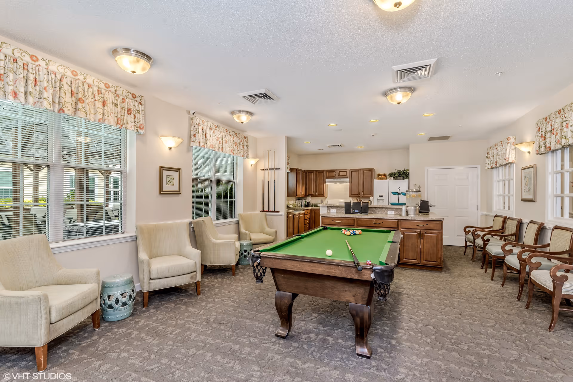 A well-lit recreational room featuring a pool table with billiard balls and cues, surrounded by beige upholstered chairs and wooden armchairs. The room has large windows with floral valances, a carpeted floor, and a kitchen area with wooden cabinets and a refrigerator in the background.