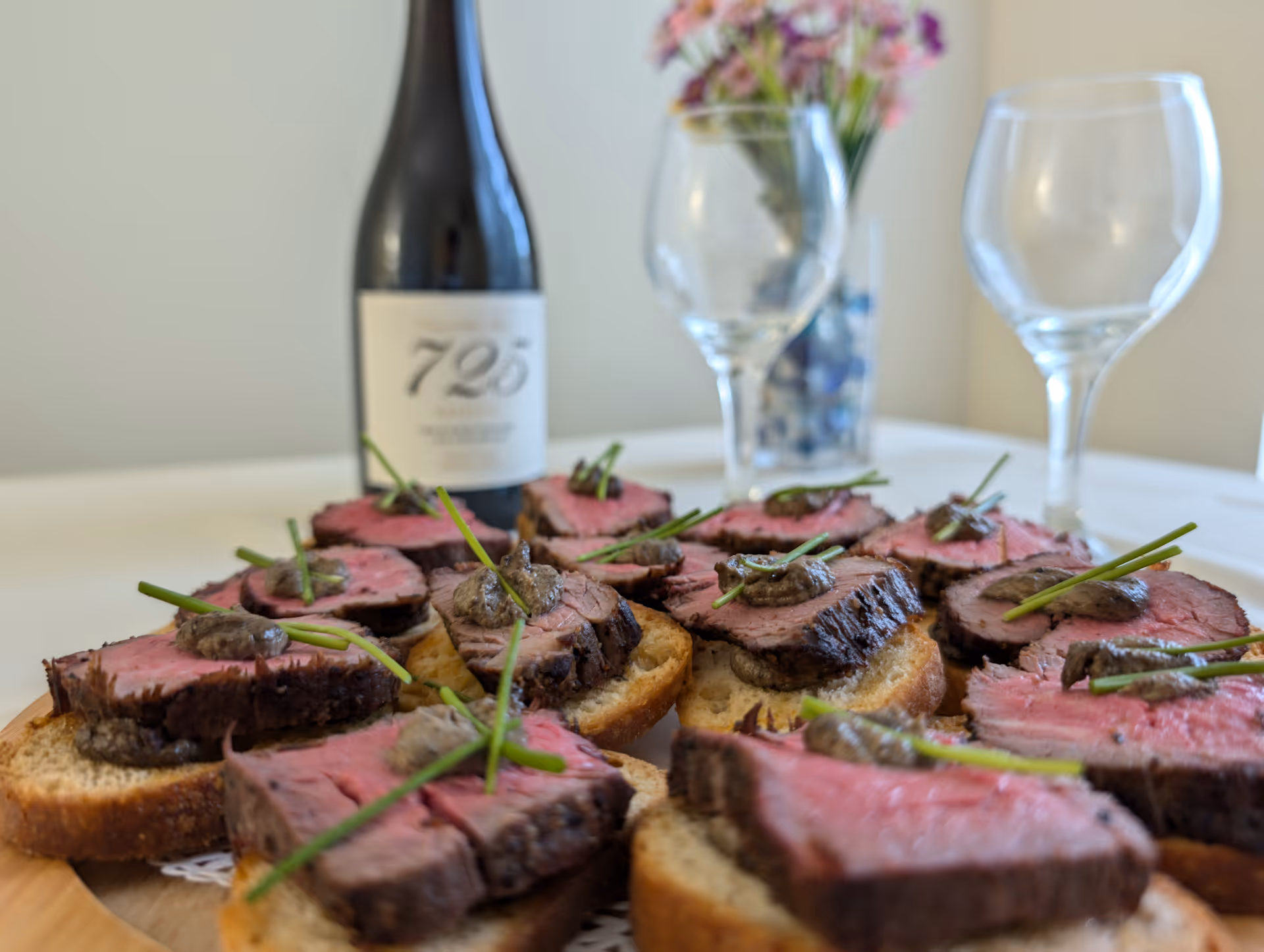 Close-up of sliced steak crostini garnished with chives on a wooden platter, with a wine bottle and two wine glasses blurred in the background.