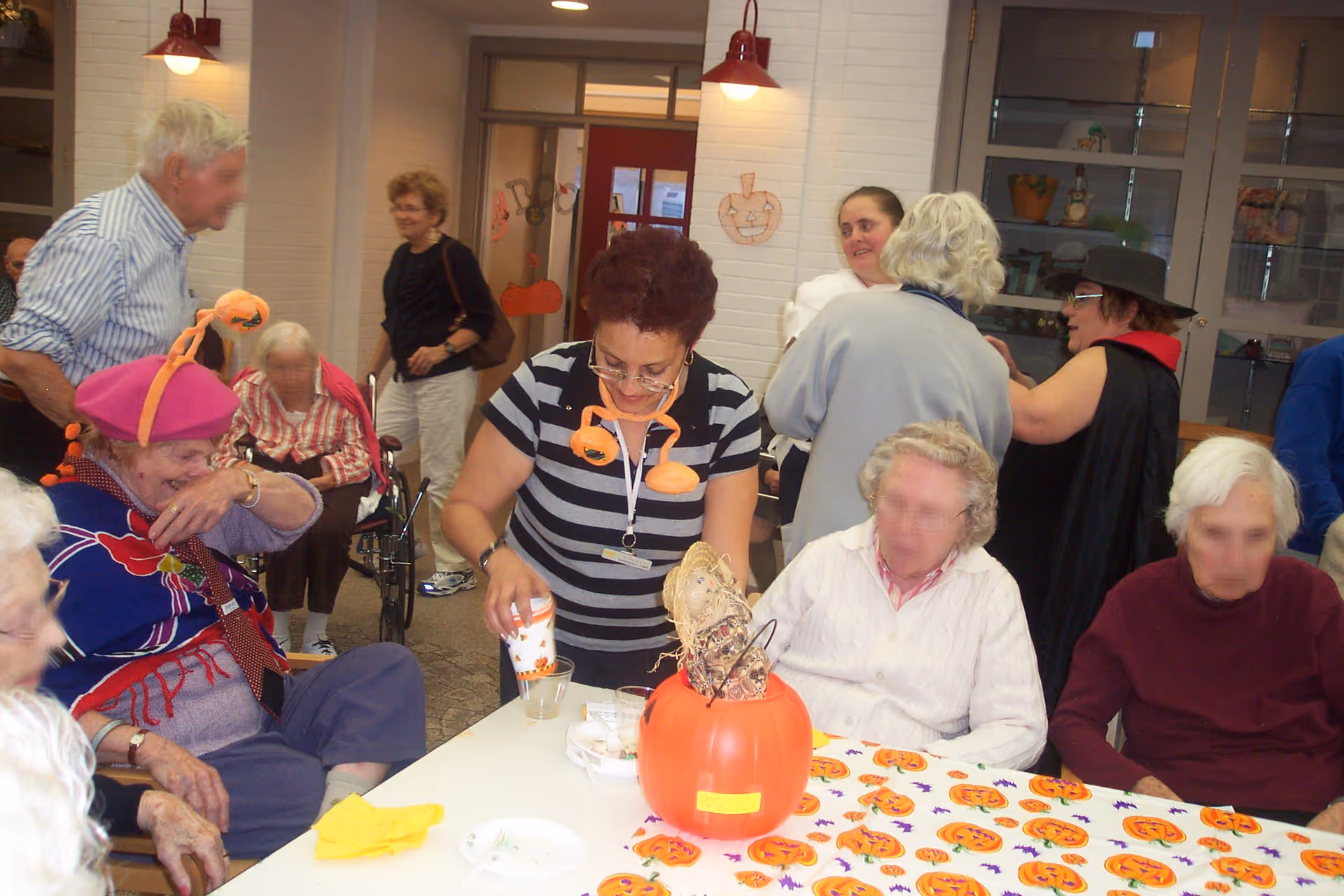A group of elderly people and a caregiver gathered around a table decorated with a Halloween-themed tablecloth and a pumpkin centerpiece. Some individuals are seated while others stand and interact in a communal indoor space with white brick walls and red light fixtures. The atmosphere appears festive and social.