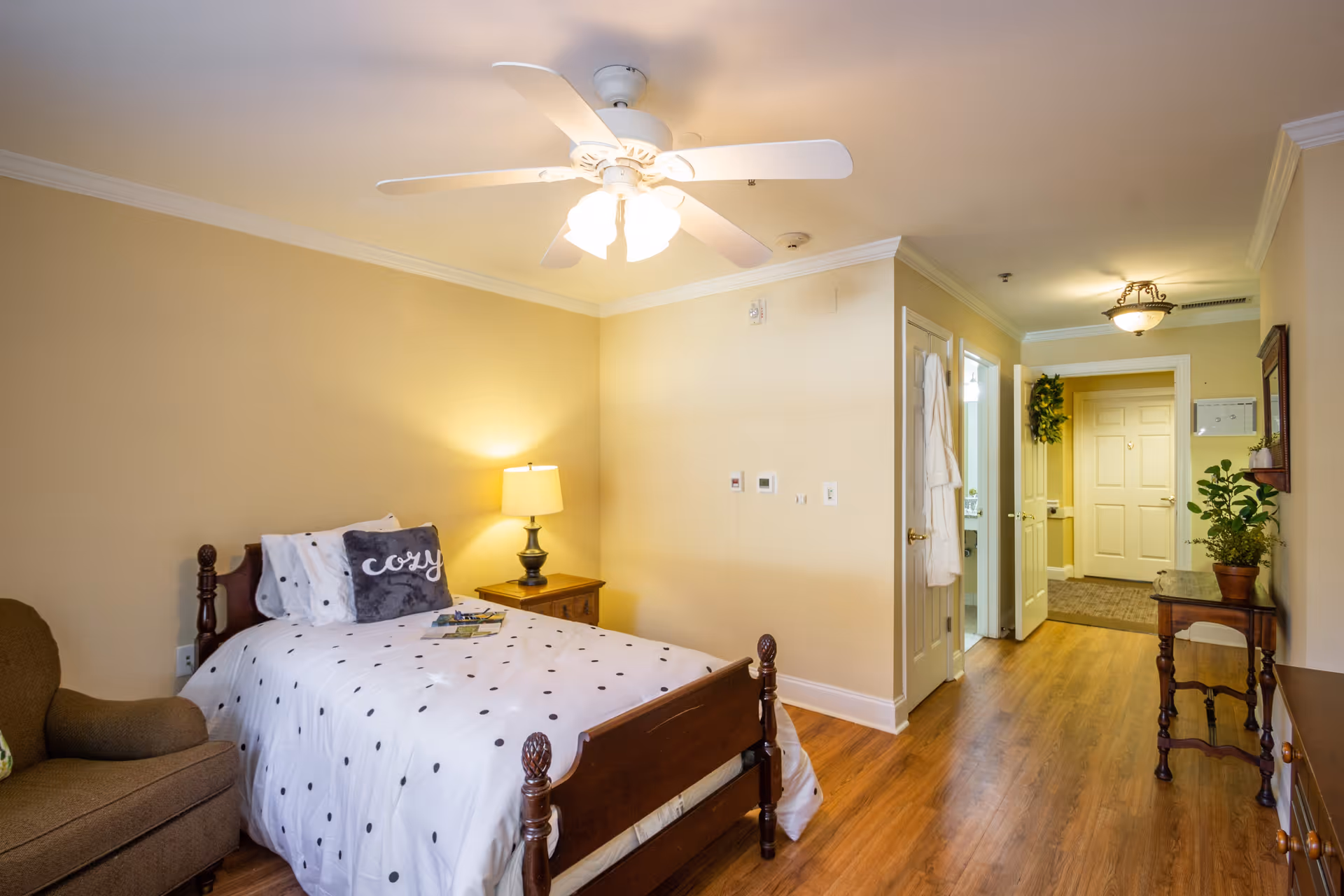 A cozy single bedroom with a bed, nightstand and lamp, ceiling fan, armchair, and a hallway leading to the entry door.