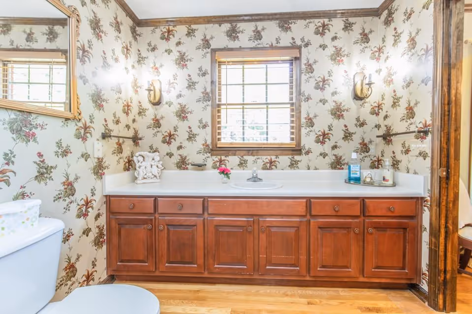 Bathroom with floral wallpaper, a wooden vanity with multiple drawers, a white countertop with a sink, a window with wooden blinds above the sink, a toilet partially visible on the left, and decorative items including a small flower vase and toiletries on the countertop.