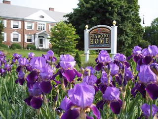 A garden with blooming purple irises in the foreground and a large brick building in the background. A sign reading 'Peabody Home Senior Living Community' is prominently displayed among the flowers.