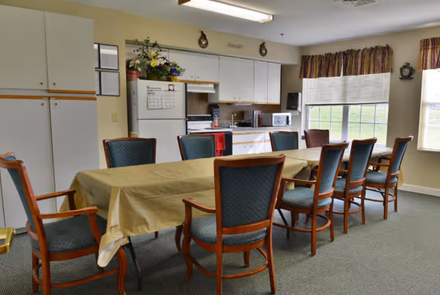 Communal dining room with a long table covered by a tablecloth surrounded by chairs, adjacent to a small kitchen with white cabinets, refrigerator, and windows.