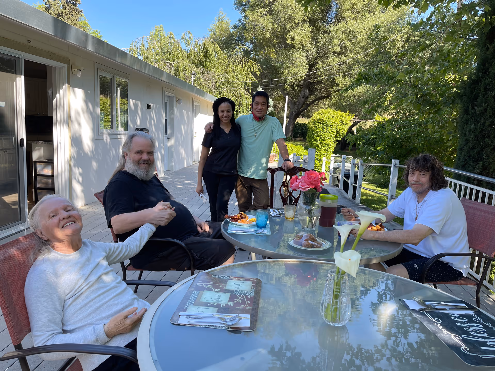 A group of five people enjoying a meal together outdoors on a patio. Three elderly individuals are seated around a glass table with plates of food and drinks, while two younger adults stand behind them smiling. The setting is surrounded by greenery and trees, with a white building in the background.