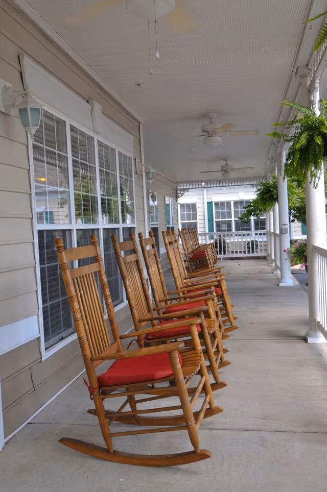A row of wooden rocking chairs with red cushions lined up on a covered porch outside a building. The porch has ceiling fans, hanging plants, and white railings, with large windows along the building wall.
