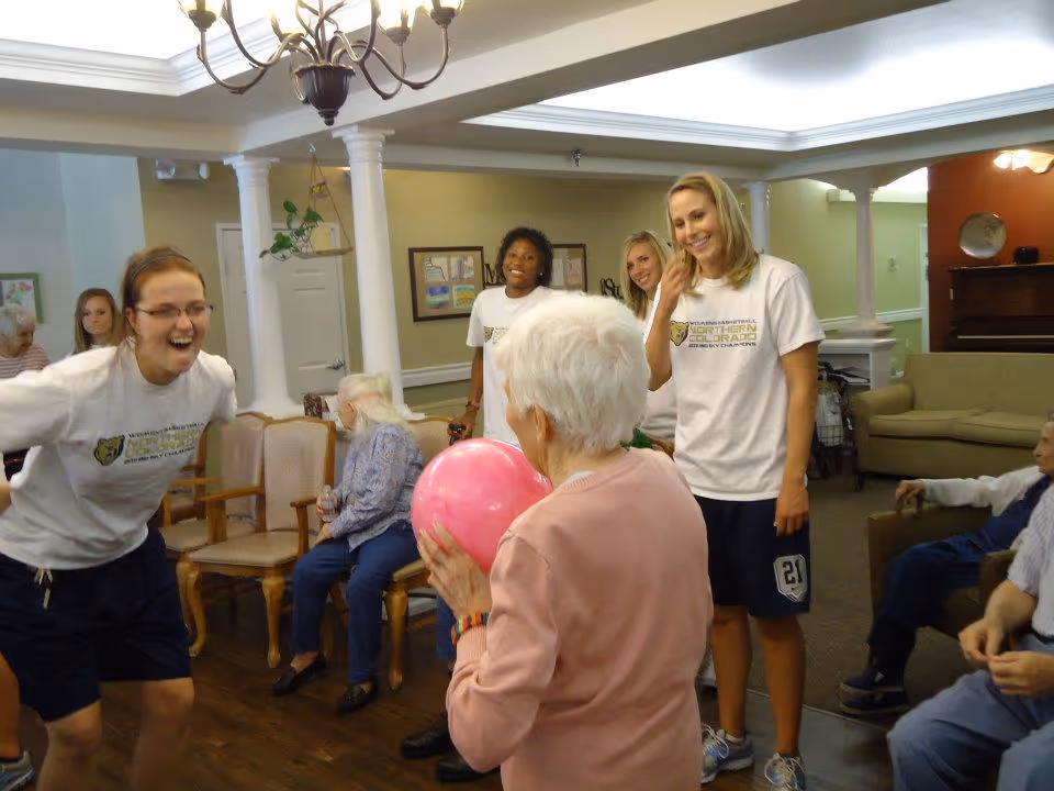 A group of elderly residents and younger adults in a common area of a senior living facility. One elderly woman is holding a pink ball while others, including three young women wearing matching white t-shirts and shorts, are smiling and interacting with her. The room has beige walls, wooden floors, chairs, and a chandelier hanging from the ceiling.