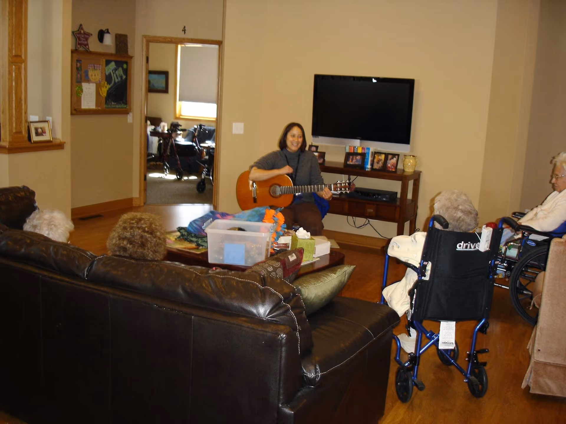 A woman playing a guitar in a living room area with several elderly people seated around her, some in wheelchairs. The room has a large flat-screen TV mounted on the wall, a wooden TV stand with framed photos, and a coffee table with various items on it. The setting appears warm and home-like.