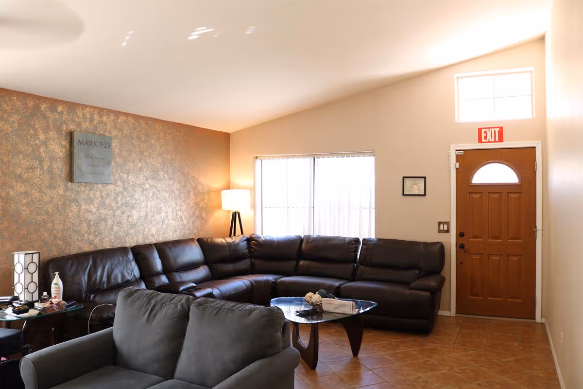 Bright living room with a large brown leather sectional, gray armchair, glass coffee table, floor lamp, and a wooden front door with an EXIT sign.