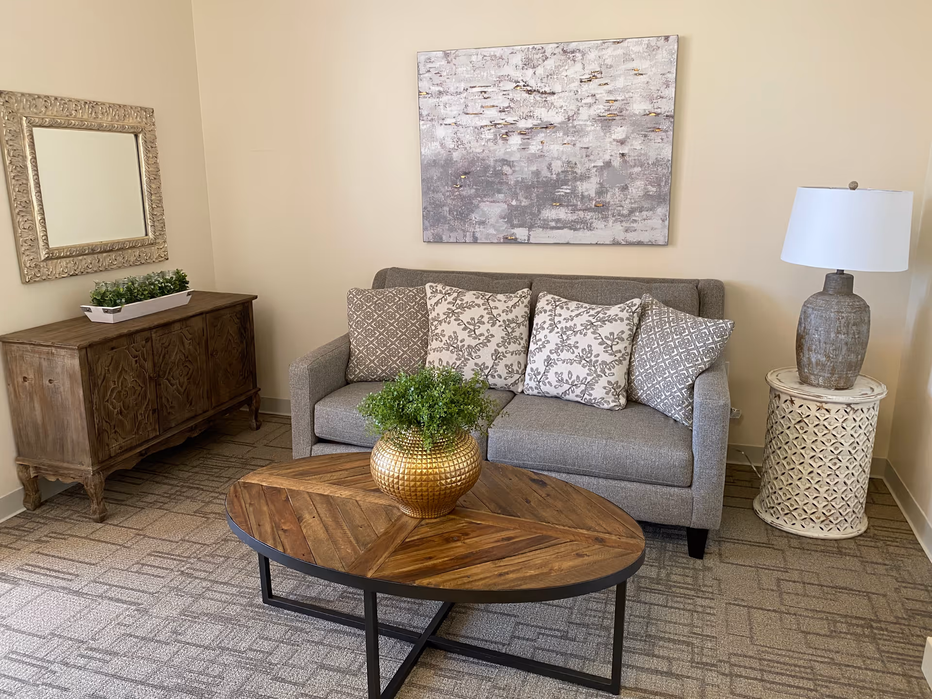 A cozy living room area with a gray sofa adorned with four patterned cushions. In front of the sofa is a wooden coffee table with a golden vase holding green plants. To the right of the sofa is a round side table with a textured lamp. On the left side of the room, there is a wooden cabinet with a rectangular mirror hanging above it. The walls are light-colored and a large abstract painting hangs above the sofa.