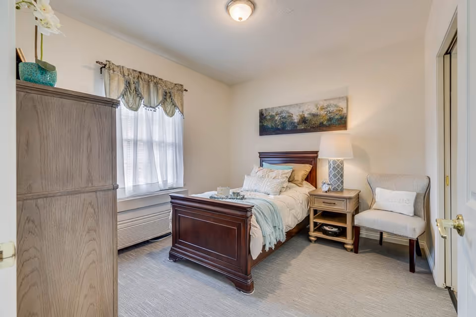 A cozy bedroom with a wooden bed frame, neatly made with white and light blue bedding and multiple pillows. Next to the bed is a wooden nightstand with a decorative lamp and a small clock. A cushioned chair with a pillow is placed beside the nightstand. A window with sheer white curtains and a decorative valance allows natural light into the room. A wooden armoire stands against the opposite wall, and a landscape painting hangs above the bed.