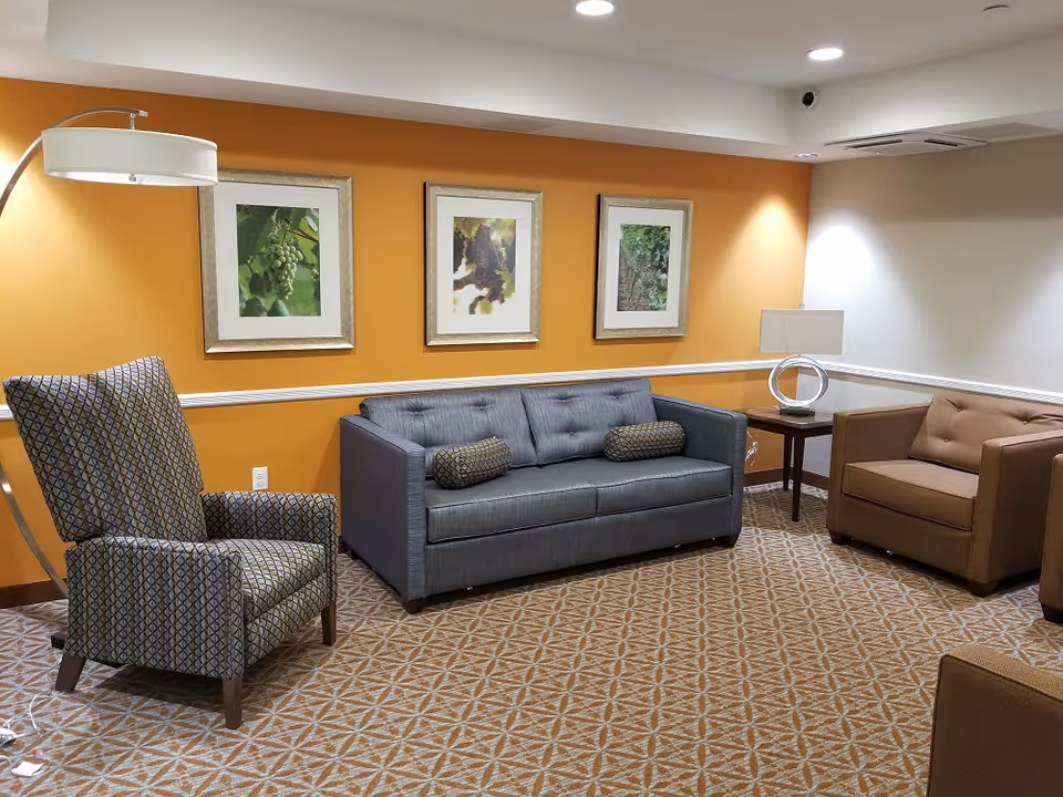 A cozy seating area with a gray sofa, patterned armchair, brown chairs, three framed pictures on an orange wall, and lamps.