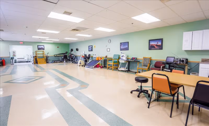 A spacious rehabilitation or physical therapy room with exercise equipment such as stationary bikes and treadmills along the green wall. There are chairs, tables, and medical devices arranged neatly. The floor has a patterned design, and the ceiling has fluorescent lighting. Several framed pictures hang on the wall.