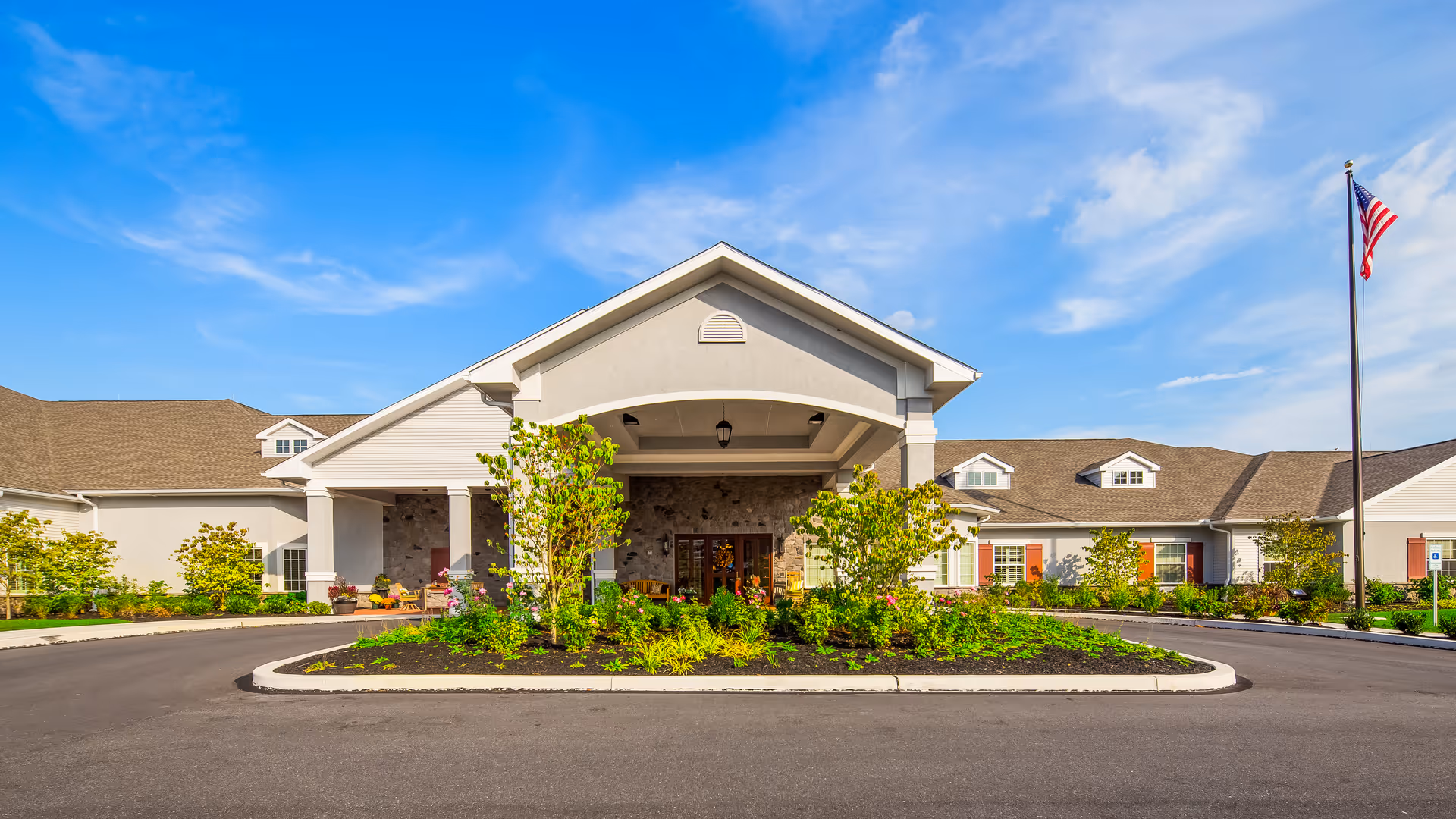 Front entrance of the Legend of Allentown senior living facility with a covered porte-cochère, landscaped island, and American flag.