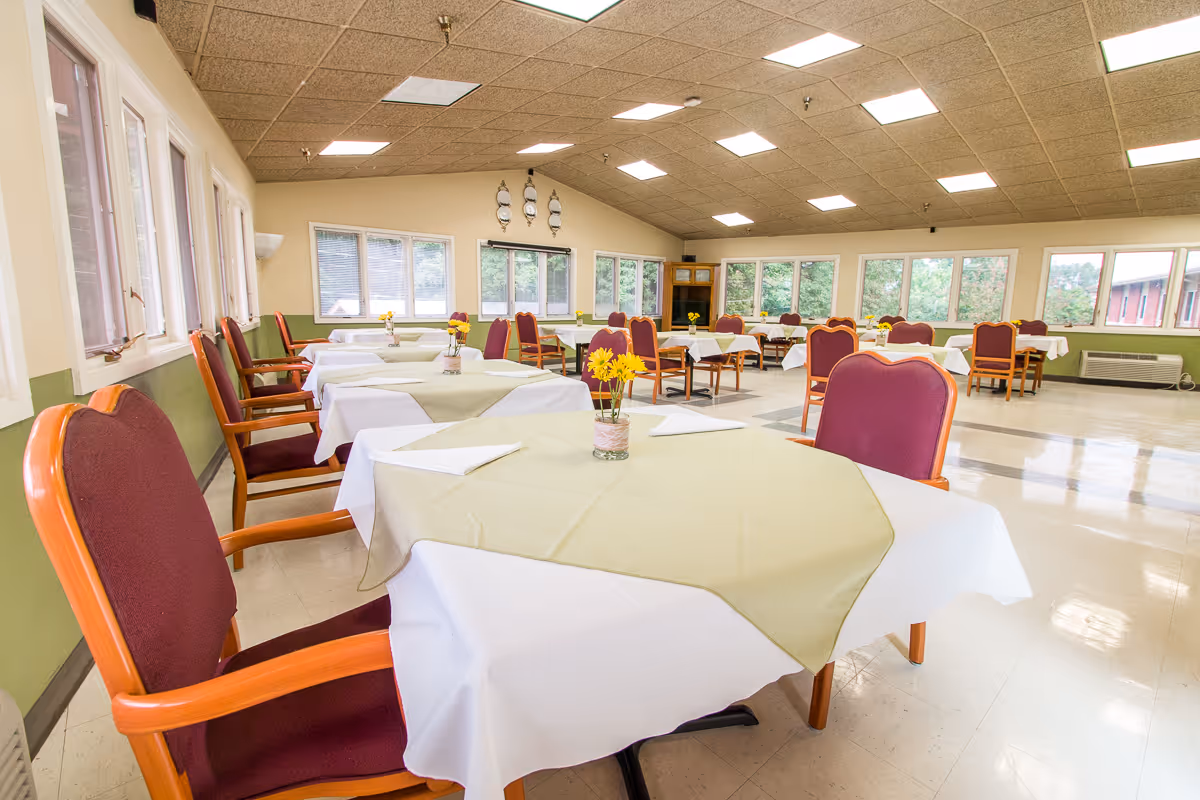 A spacious dining room with multiple tables covered in white tablecloths and beige overlays, each table decorated with a small vase of yellow flowers. The room has maroon cushioned chairs with wooden armrests, large windows letting in natural light, and a tiled floor with a green and beige color scheme.