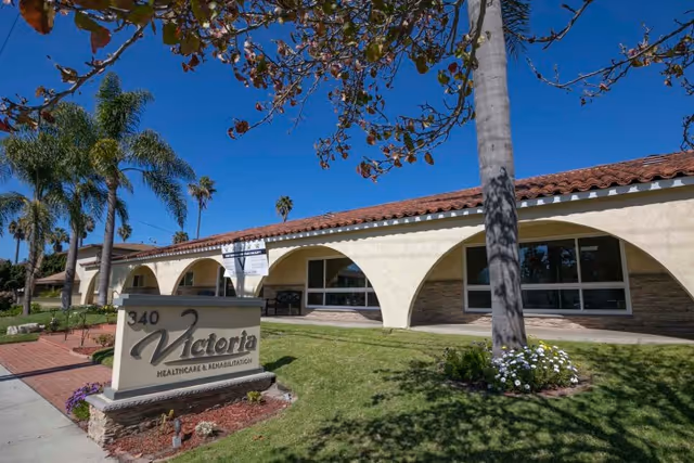 Front exterior of Victoria Healthcare & Rehabilitation with a stone sign, arched colonnade, palm trees, and a manicured lawn under a clear blue sky.