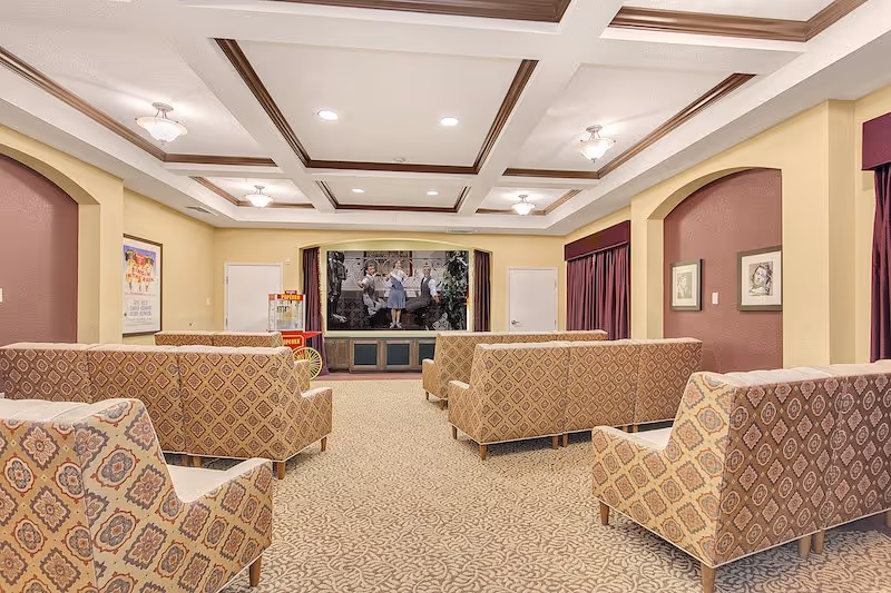 A cozy community room with patterned armchairs arranged in rows facing a small stage with a screen showing a black and white movie scene. The room has a beige carpet, cream-colored walls with maroon accents, framed pictures on the walls, and a coffered ceiling with recessed lighting.