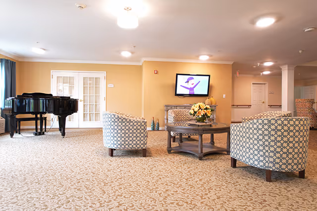A spacious senior living common area with patterned carpet, three upholstered armchairs arranged around a round wooden coffee table with a flower arrangement, a grand piano near a set of French doors, and a wall-mounted TV displaying a cartoon character.