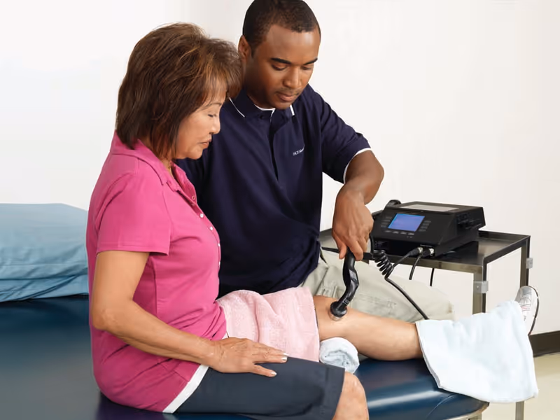 A healthcare professional using a therapeutic ultrasound device on a woman's knee while she sits on a treatment table in a clinical setting.