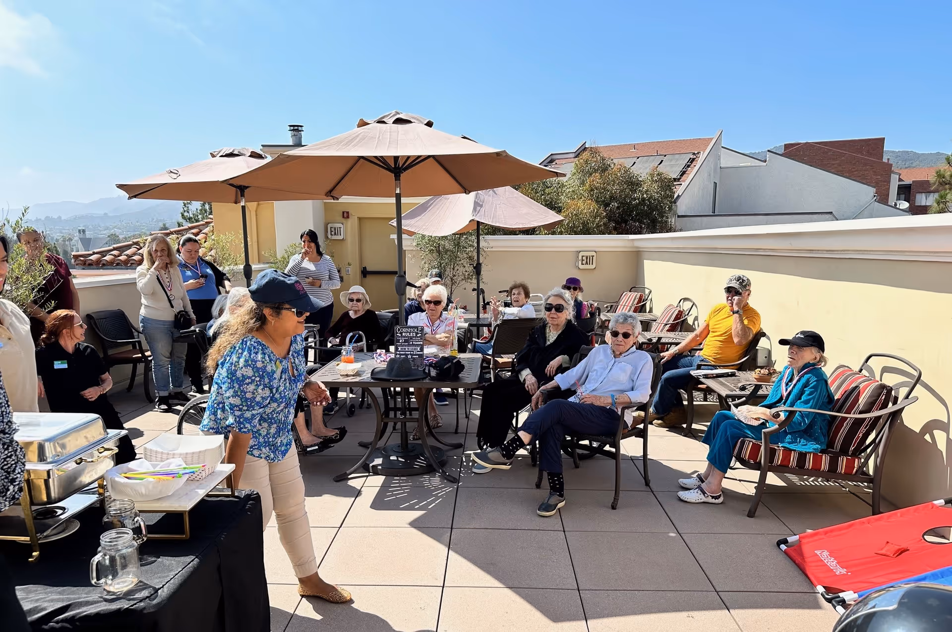 A group of elderly people sitting and socializing on an outdoor patio with tables, chairs, and large umbrellas providing shade. Some people are seated while others are standing, enjoying a sunny day with a view of distant hills and buildings.